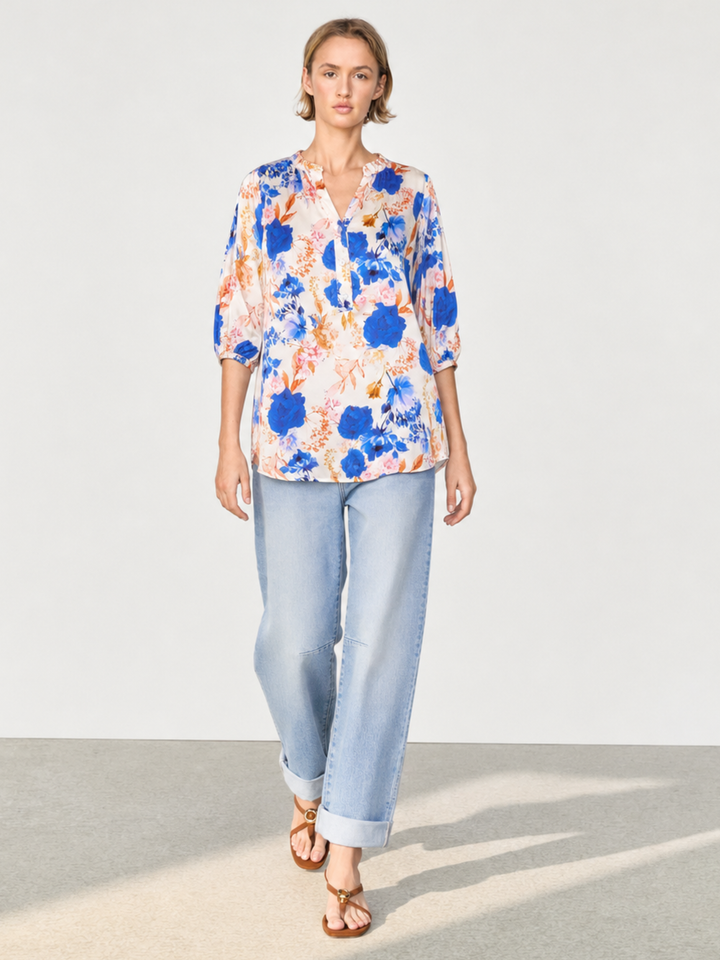 A woman modeling a short-sleeve button-up blouse with a blue, orange, and white abstract floral print, paired with light-wash cuffed jeans and brown sandals, set against a minimalist studio background.