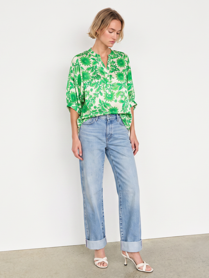 A woman modeling a green and white tropical floral print blouse with light-wash blue jeans and white heeled sandals, standing against a plain white wall.