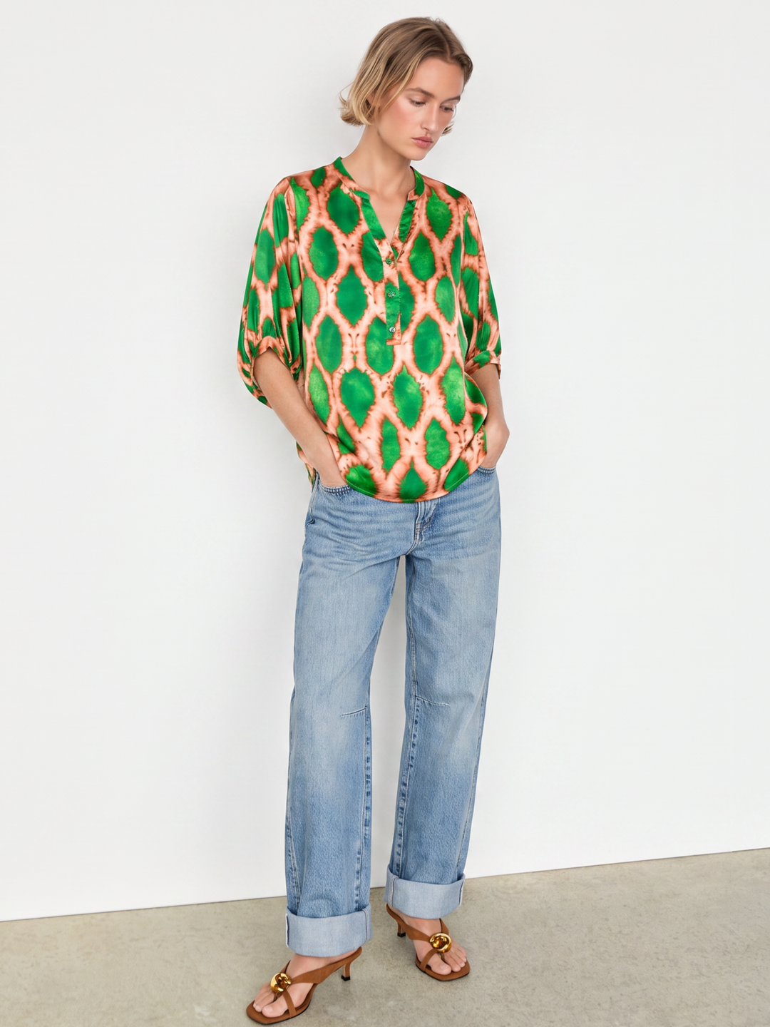 A woman modeling a vibrant green and orange patterned blouse with three-quarter sleeves, paired with light-wash blue jeans and brown sandals, standing against a plain white wall.