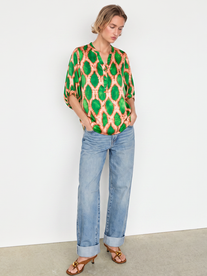 A woman modeling a vibrant green and orange patterned blouse with three-quarter sleeves, paired with light-wash blue jeans and brown sandals, standing against a plain white wall.