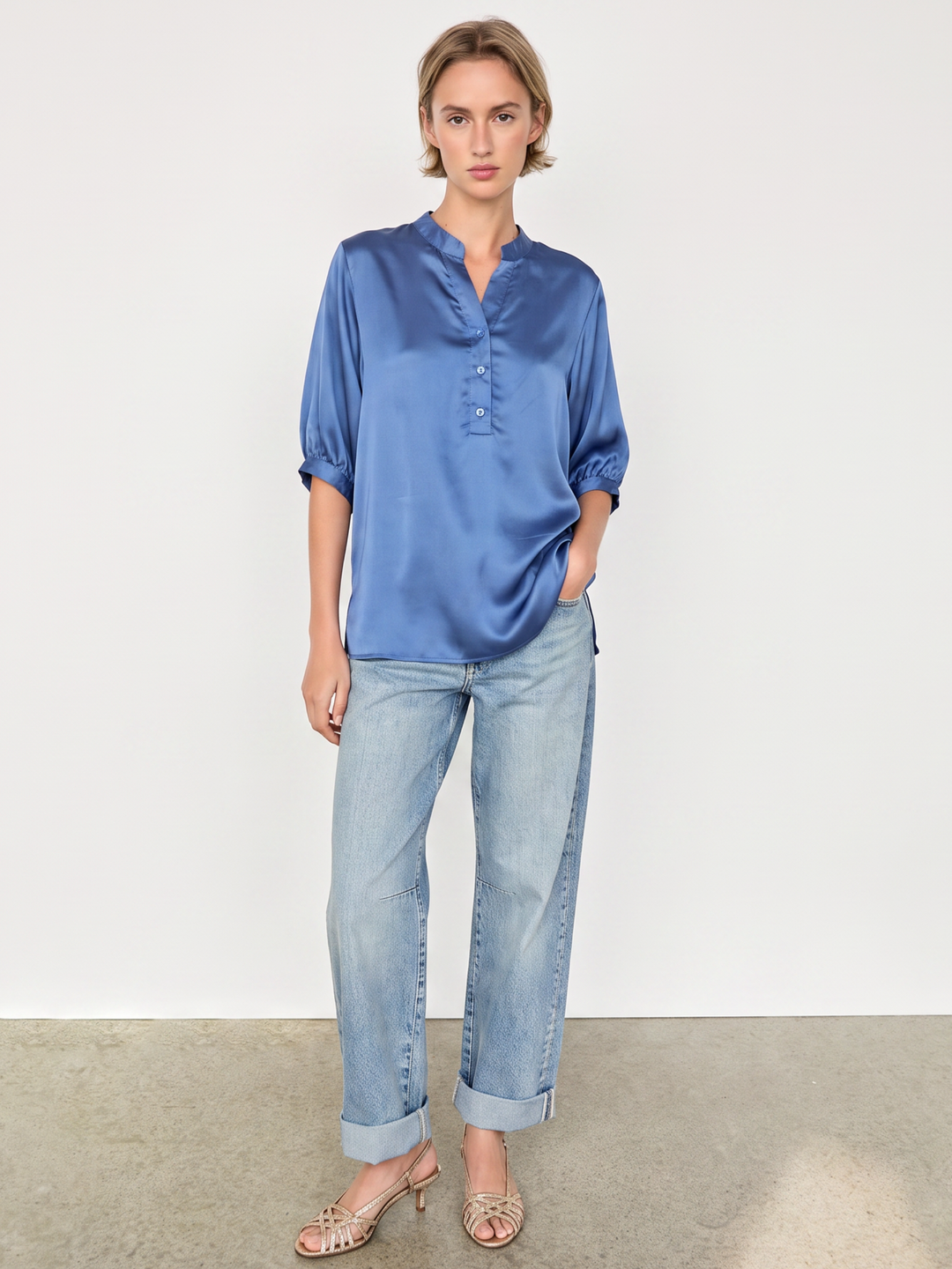 A woman modeling a blue satin blouse with three-quarter sleeves and a buttoned stand collar, paired with light-wash blue jeans and beige heeled sandals, standing against a plain off-white wall.