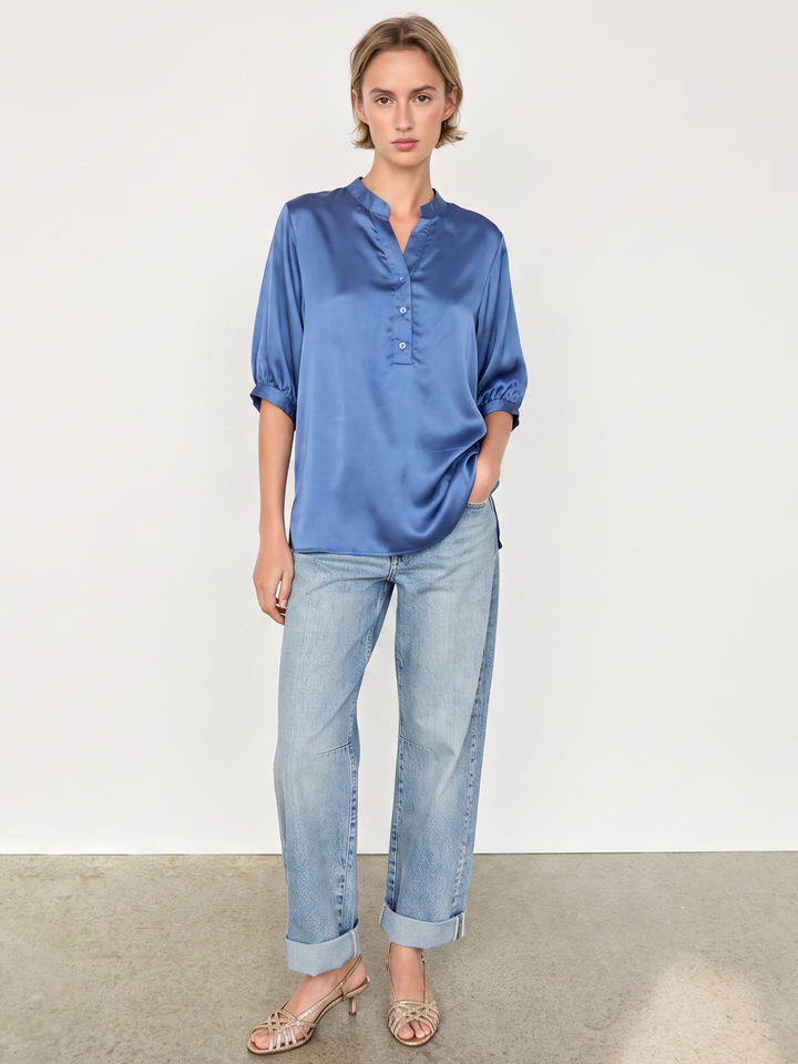 A woman modeling a blue satin blouse with three-quarter sleeves and a buttoned stand collar, paired with light-wash blue jeans and beige heeled sandals, standing against a plain off-white wall.