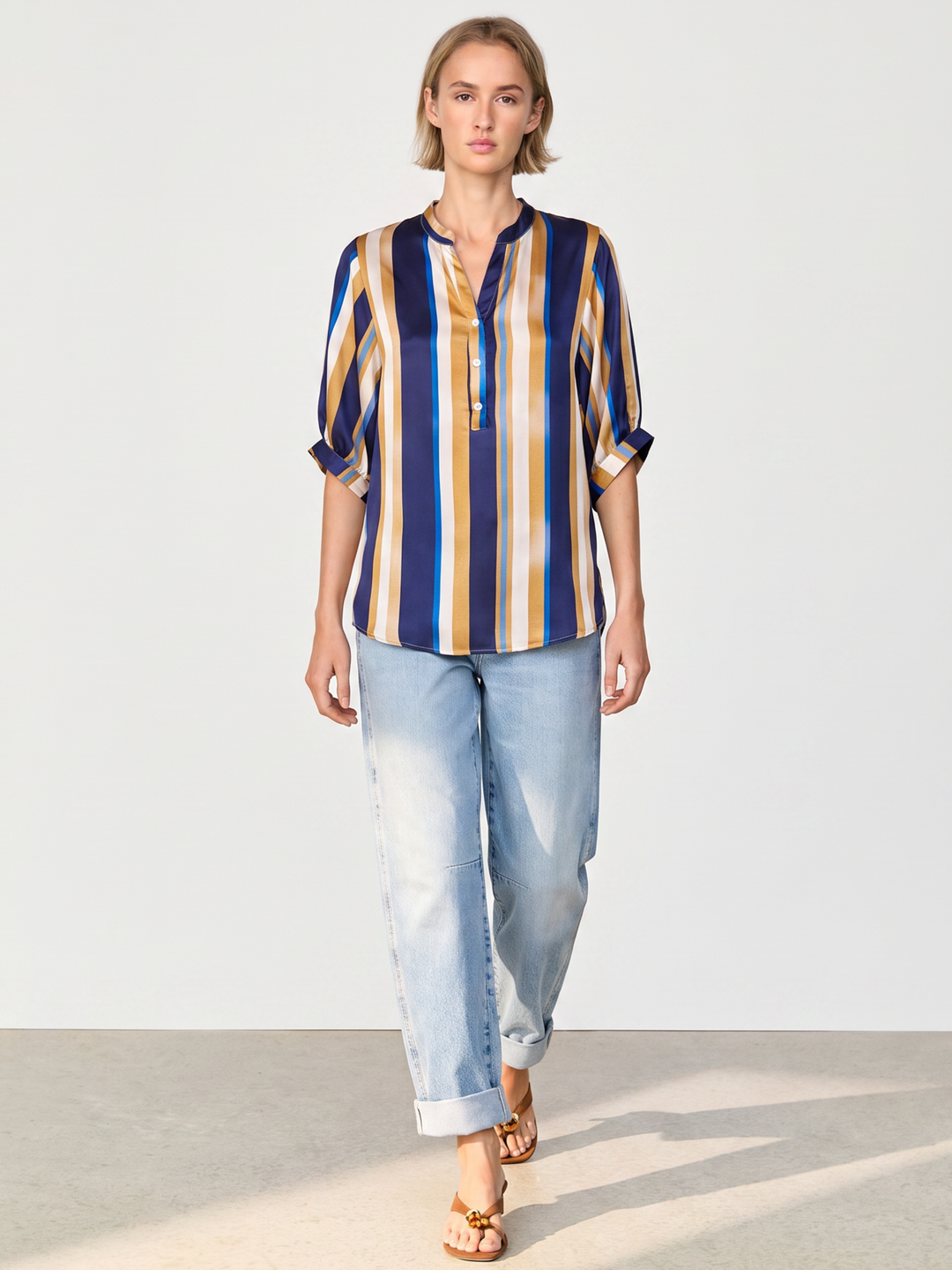 A woman modeling a short-sleeve button-front blouse with vertical navy, mustard yellow, and beige stripes, paired with light-wash blue jeans and tan sandals, against a plain light gray studio background.