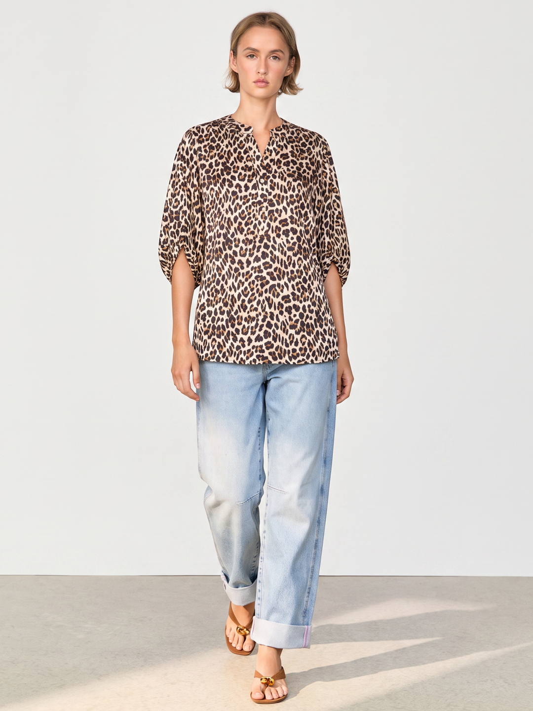 A woman modeling a leopard-print blouse with three-quarter sleeves and light-wash blue jeans, standing against a plain light gray studio background.