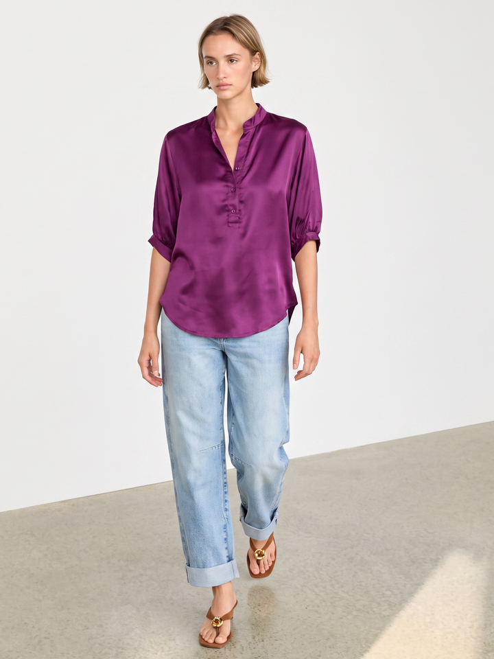 A woman modeling a purple satin button-up blouse with rolled sleeves, paired with light-wash blue jeans and gold sandals, standing in a minimalist studio setting.