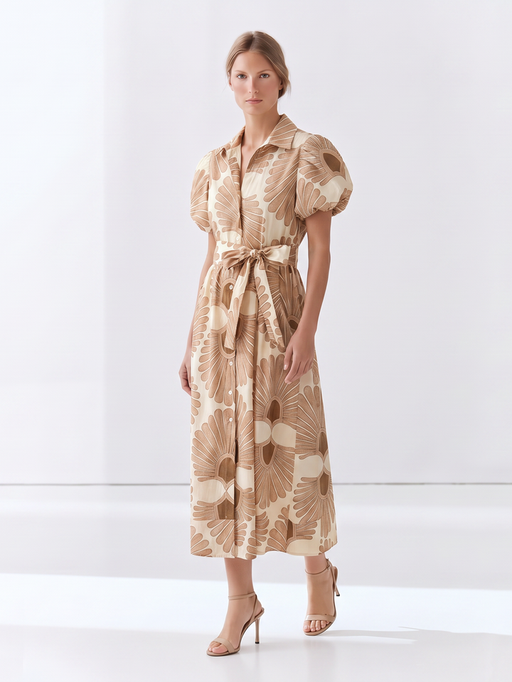A woman modeling a midi-length beige and brown floral print dress with puff sleeves, a pointed collar, and a matching waist tie, paired with nude high-heeled sandals in a bright studio setting.