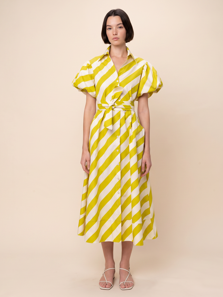 A woman modeling a yellow and white diagonal striped midi dress with puff sleeves and a matching tie belt, set against a solid beige studio background.