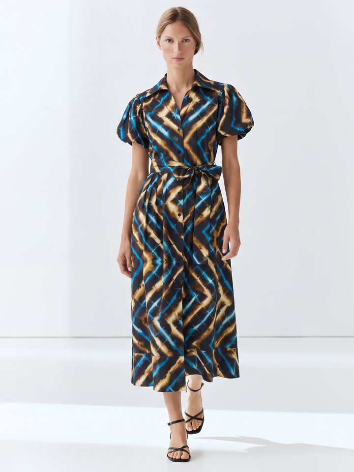 A woman modeling a short-sleeve midi shirt dress with a geometric zigzag pattern in blue, brown, and beige, featuring a tie waist and collar, set against a plain white studio background.