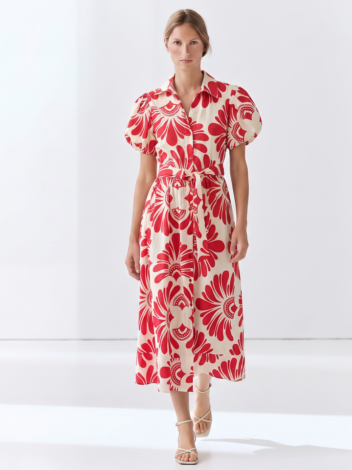 A woman modeling a midi-length cream dress with a bold red floral pattern, featuring puff sleeves and a matching tie belt, set against a clean white studio background.