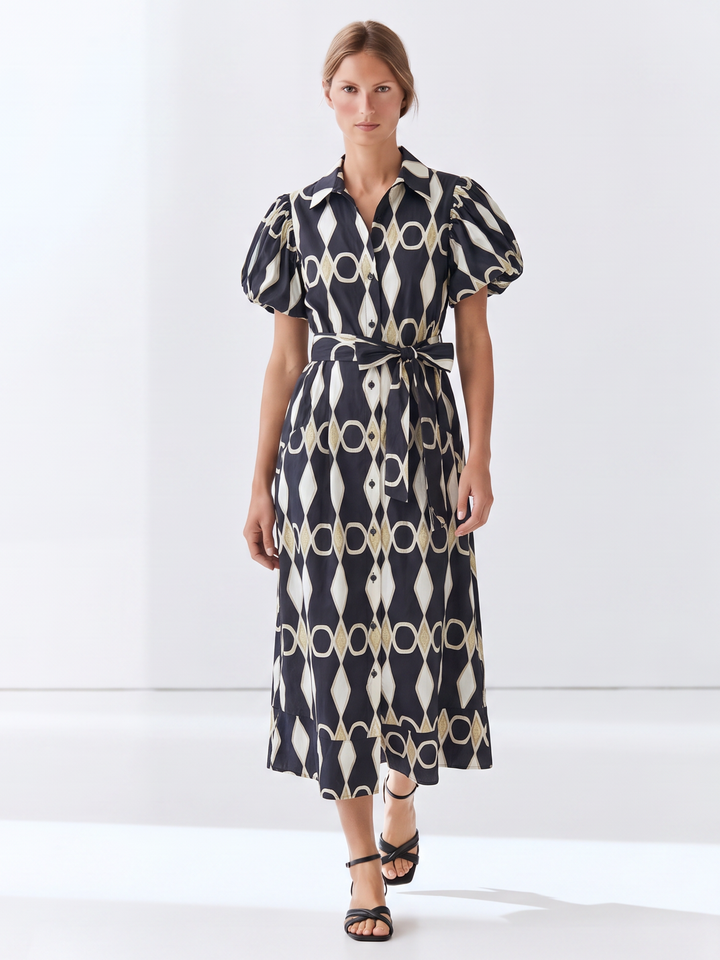 A woman modeling a midi shirt dress with puff sleeves, a self-tie waist belt, and an abstract geometric chain-link pattern in black, cream, and beige, set against a clean white studio background.