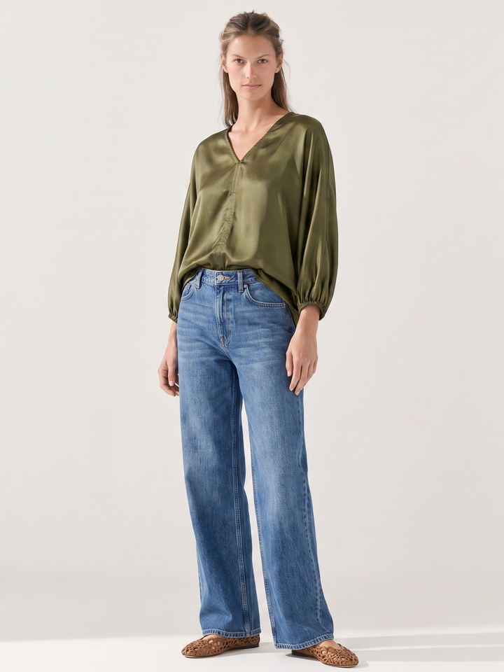 A woman modeling a sage green satin long-sleeve blouse with balloon sleeves and blue wide-leg jeans, standing against a plain light gray studio background.