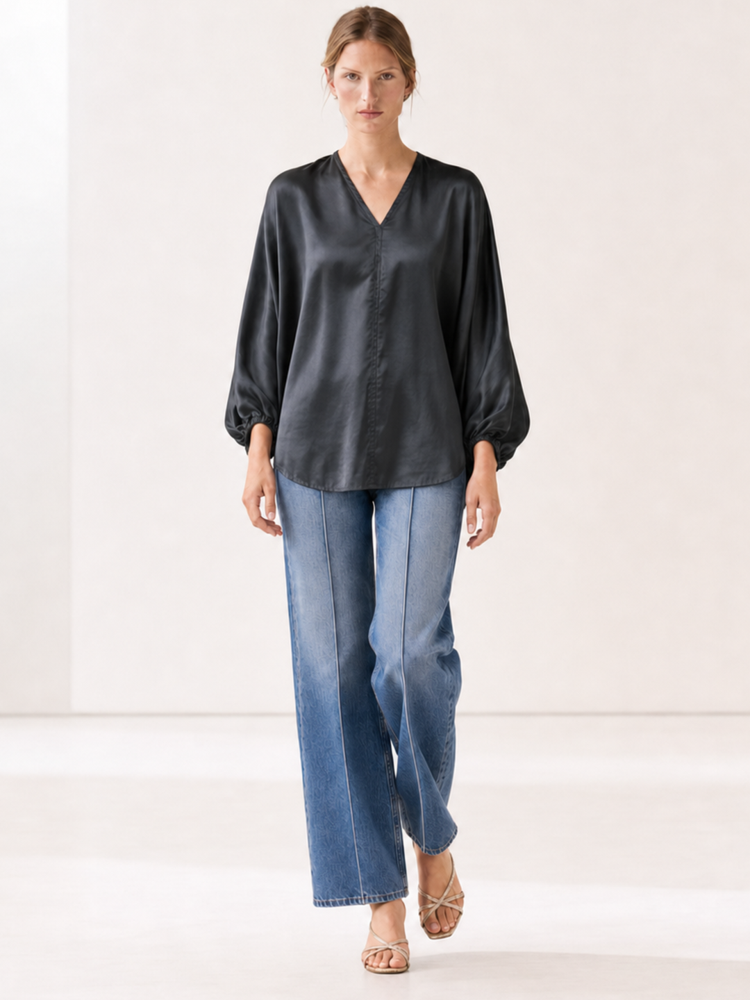 A woman modeling a black satin-like long-sleeve blouse with a V-neck and wide cuffs, paired with blue jeans and beige sandals, against a plain light studio background.
