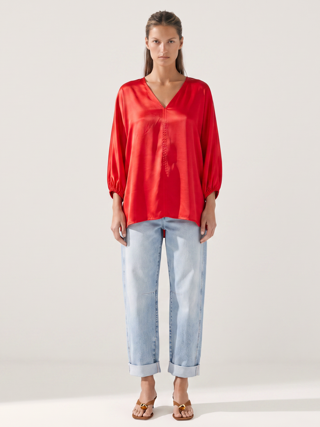 A woman modeling a vibrant red satin long-sleeve blouse with a V-neck and button-down front, paired with light-wash blue jeans and brown sandals, against a plain light gray studio background.