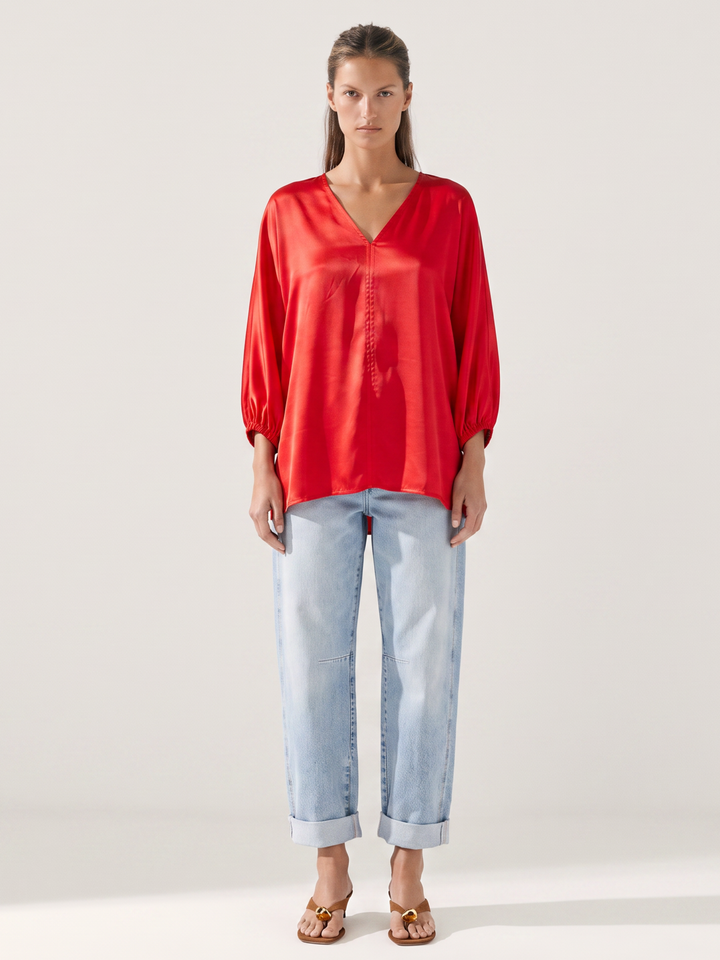 A woman modeling a vibrant red satin long-sleeve blouse with a V-neck and button-down front, paired with light-wash blue jeans and brown sandals, against a plain light gray studio background.