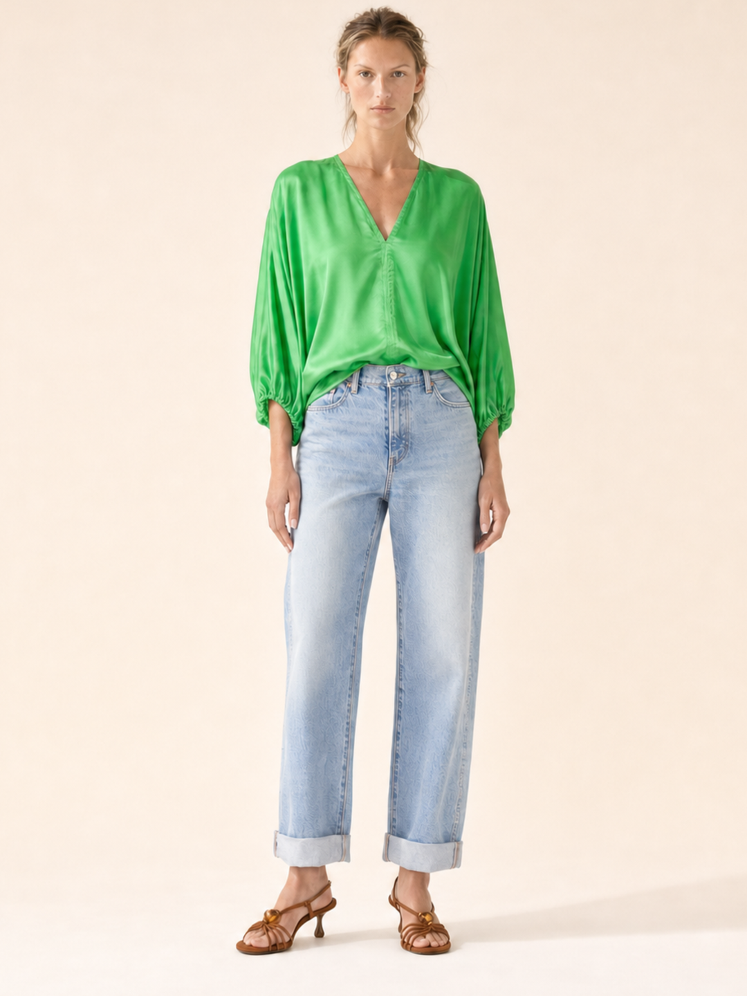 A woman modeling a vibrant green satin blouse with voluminous three-quarter sleeves, paired with light-wash blue jeans and brown strappy sandals, set against a solid warm beige background.