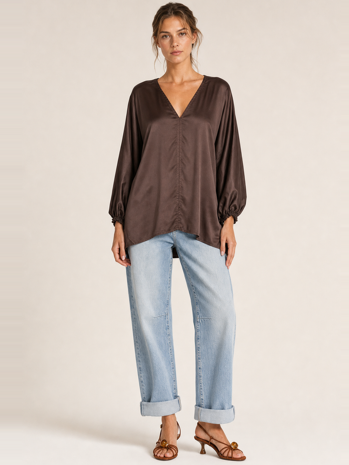 A woman modeling a brown satin long-sleeve blouse with a gathered front and balloon sleeves, paired with light blue jeans and brown heeled sandals, against a plain beige studio background.