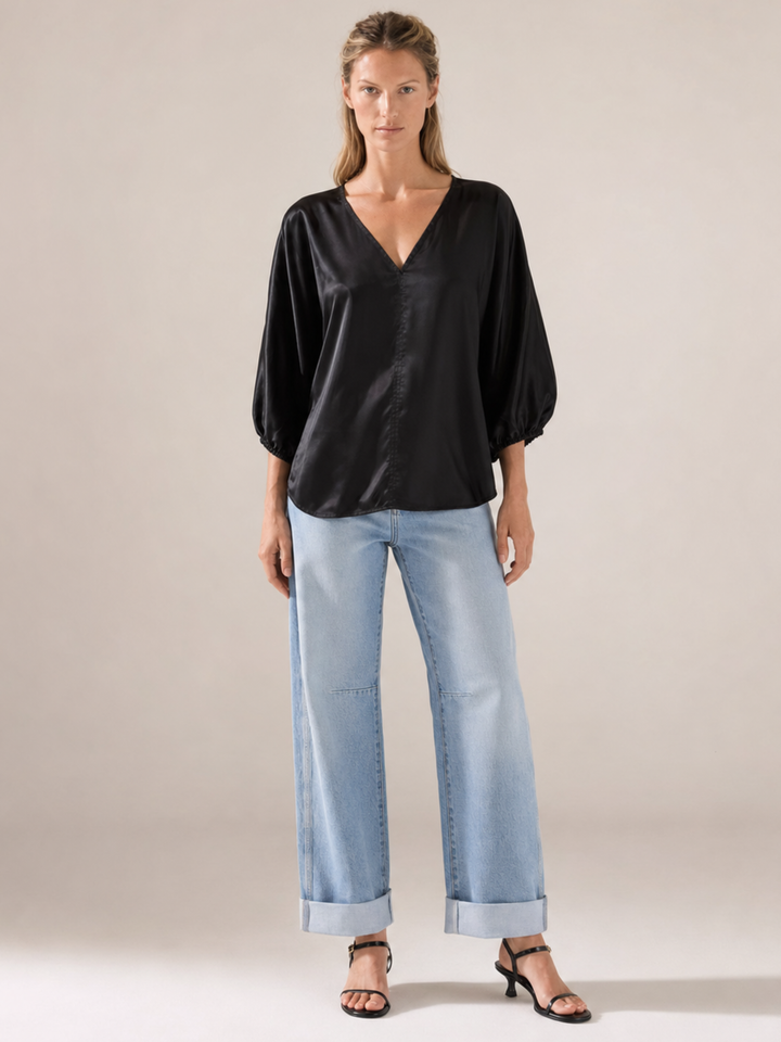 A woman modeling a black satin-like V-neck blouse with voluminous elbow-length sleeves, paired with light-wash wide-leg jeans and black heeled sandals, against a plain warm beige studio background.