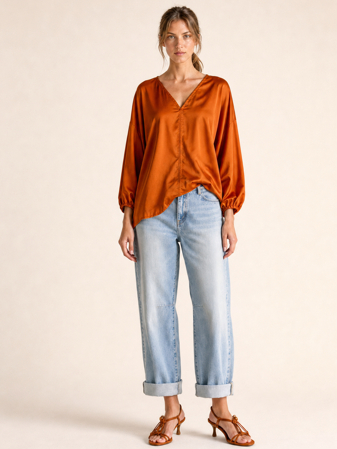 A woman modeling a long-sleeve, burnt orange satin blouse with a V-neck and gathered cuffs, paired with light-wash cuffed jeans and brown strappy sandals, against a solid beige studio background.