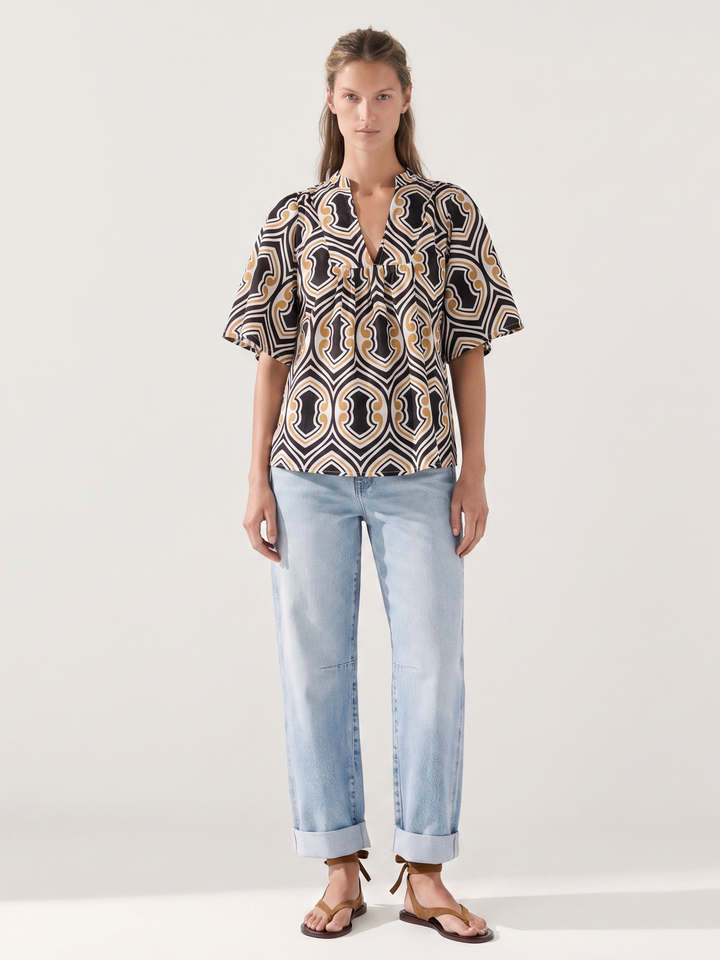 A woman modeling a short-sleeve button-up shirt with a black, beige, and gold geometric pattern, paired with light-wash blue jeans and brown sandals, against a plain light gray studio background.