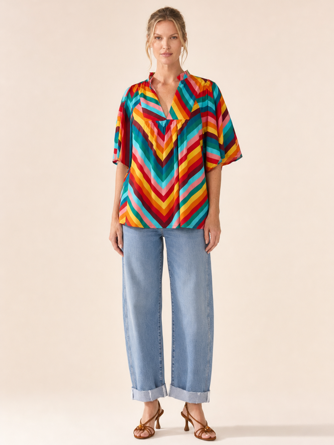 A woman modeling a short-sleeve button-up blouse with a bold multicolor chevron pattern in red, orange, yellow, green, blue, and purple, paired with light-wash wide-leg jeans and brown heeled sandals, against a solid beige studio background.