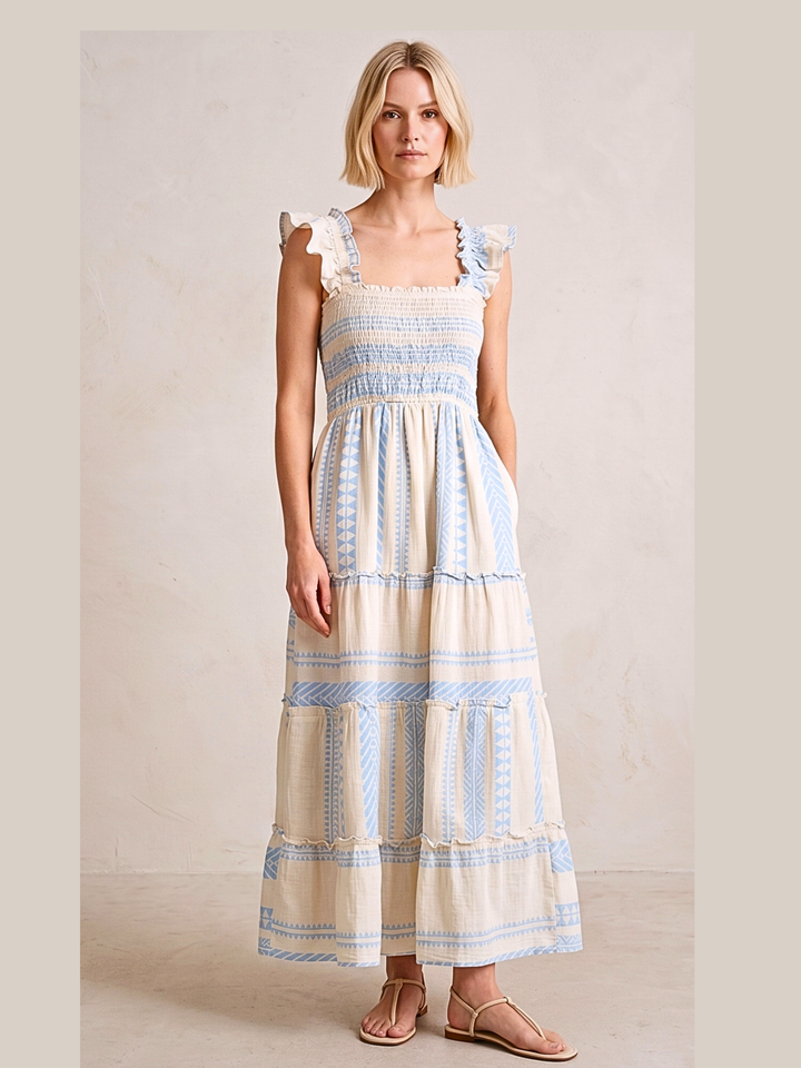 A woman modeling a tiered maxi dress with a cream base and light blue geometric stripes, featuring ruffled shoulder straps and side pockets, set against a neutral beige background.