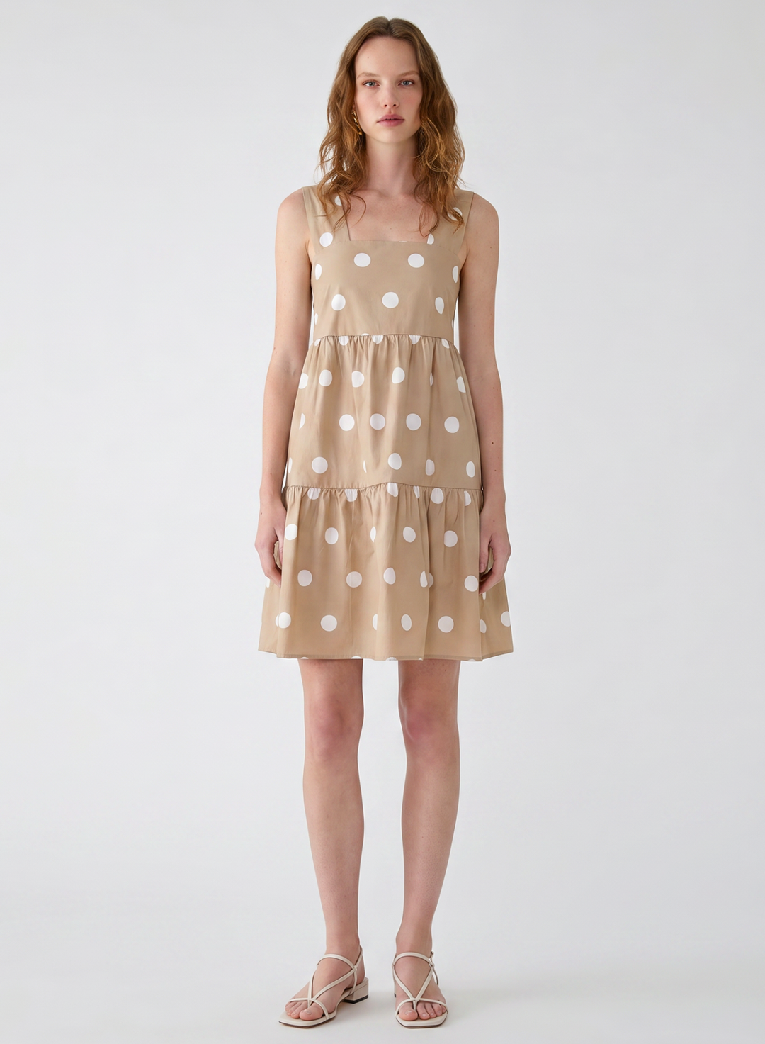 A woman modeling a sleeveless tiered mini dress in beige with a white polka dot pattern, paired with strappy flat sandals, against a plain light gray studio background.