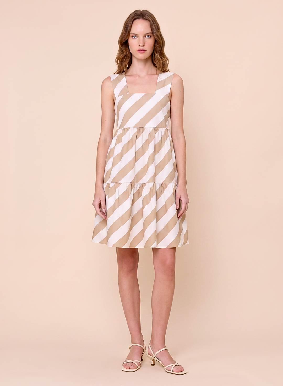 A woman modeling a sleeveless tiered dress with diagonal beige and white stripes, paired with light beige strappy sandals, set against a solid pale peach studio background.
