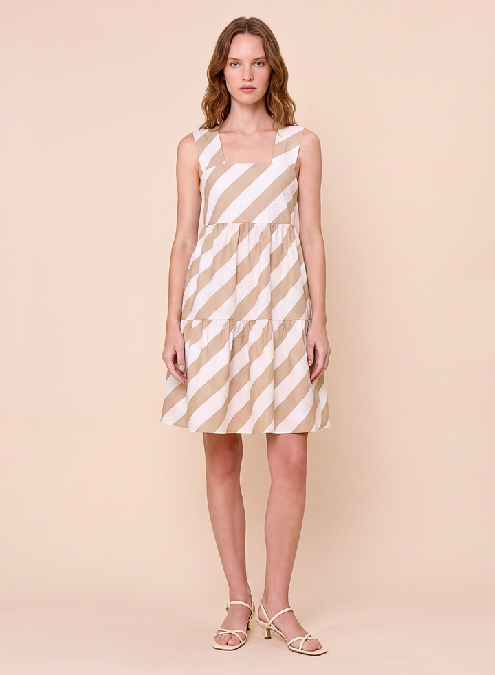 A woman modeling a sleeveless tiered dress with diagonal beige and white stripes, paired with light beige strappy sandals, set against a solid pale peach studio background.
