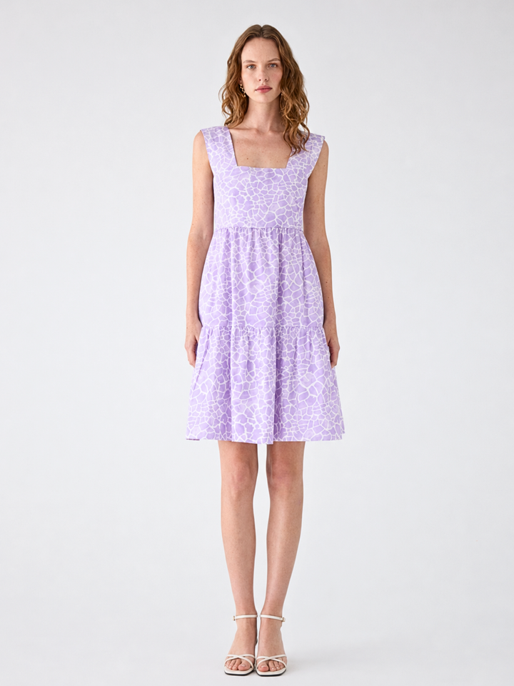 A woman modeling a sleeveless lavender dress with a subtle tonal floral pattern, a square neckline, and a tiered skirt, paired with white strappy heels against a light gray studio background.