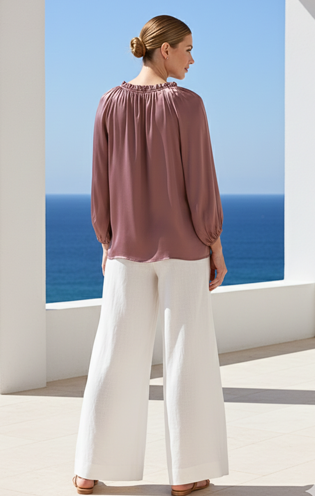 Woman wearing a mauve blouse and white pants standing in front of a scenic ocean view.