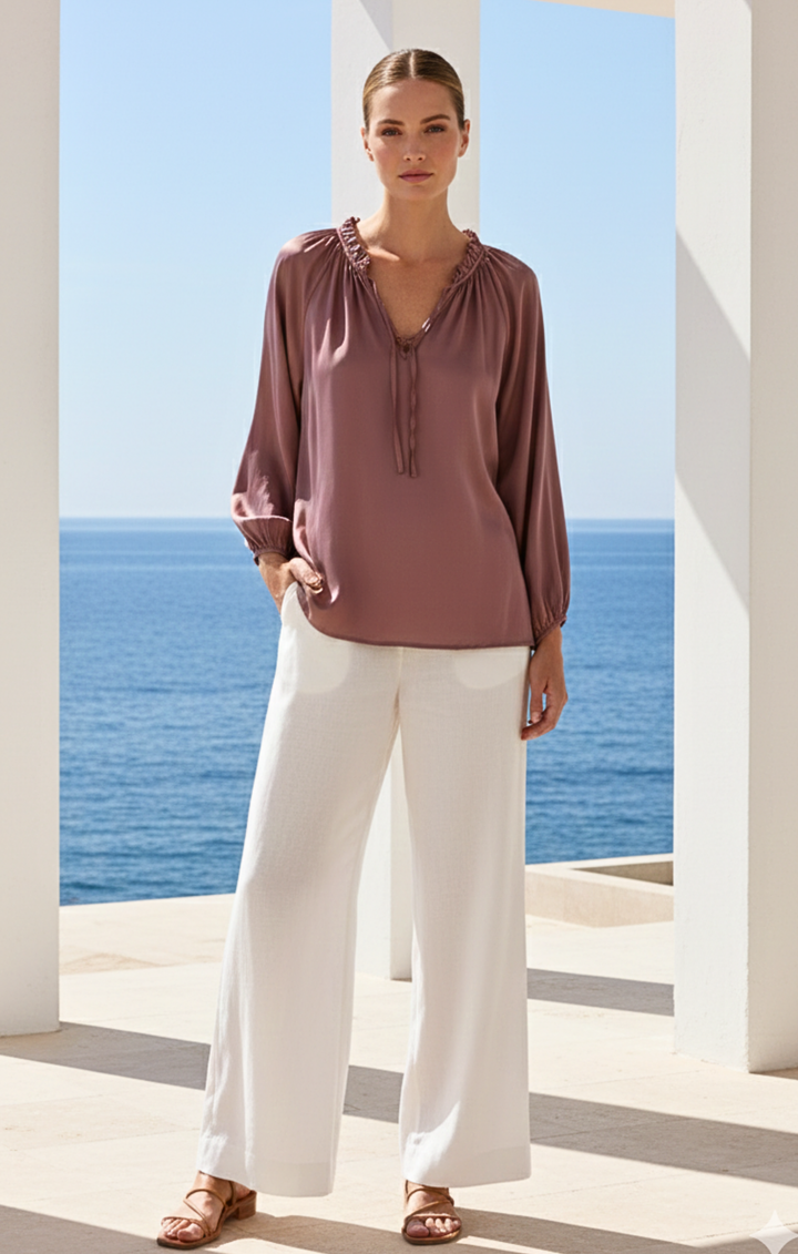 Woman wearing a mauve blouse and white pants standing in front of a scenic ocean view.