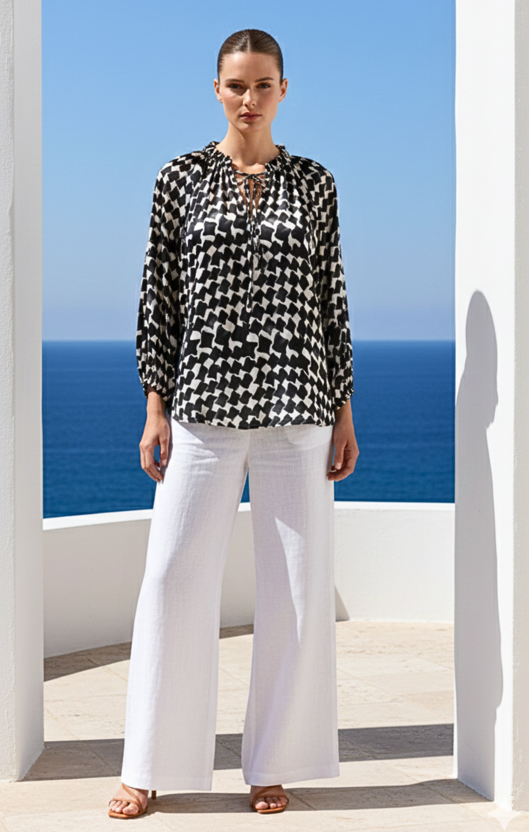Woman wearing a black and white patterned blouse and white pants standing in front of a blue ocean view.