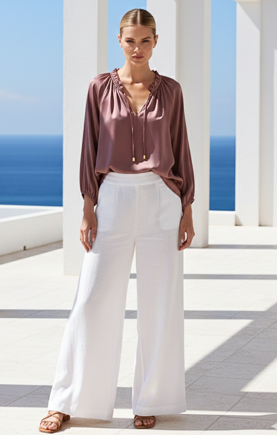 Woman wearing a mauve blouse and white pants standing in front of a blue ocean and white columns.