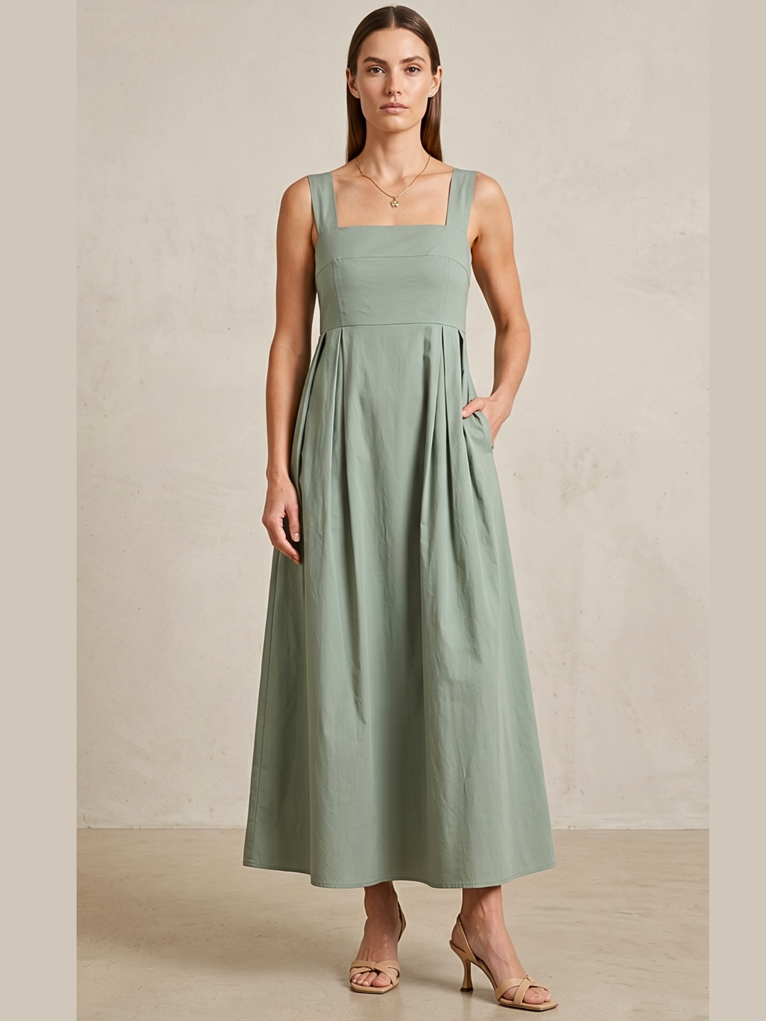 A woman modeling a sage green sleeveless maxi dress with a square neckline, structured bodice, and side pockets, standing against a neutral textured beige wall.
