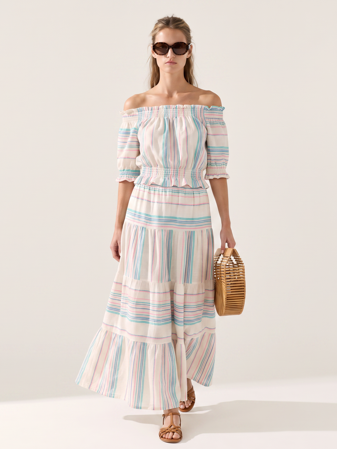 A woman modeling an off-the-shoulder maxi dress with pastel vertical stripes in pink, blue, and green on a cream background, holding a small woven straw bag against a plain light beige studio backdrop.