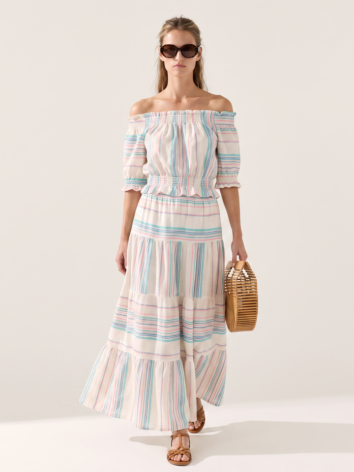 A woman modeling an off-the-shoulder maxi dress with pastel vertical stripes in pink, blue, and green on a cream background, holding a small woven straw bag against a plain light beige studio backdrop.
