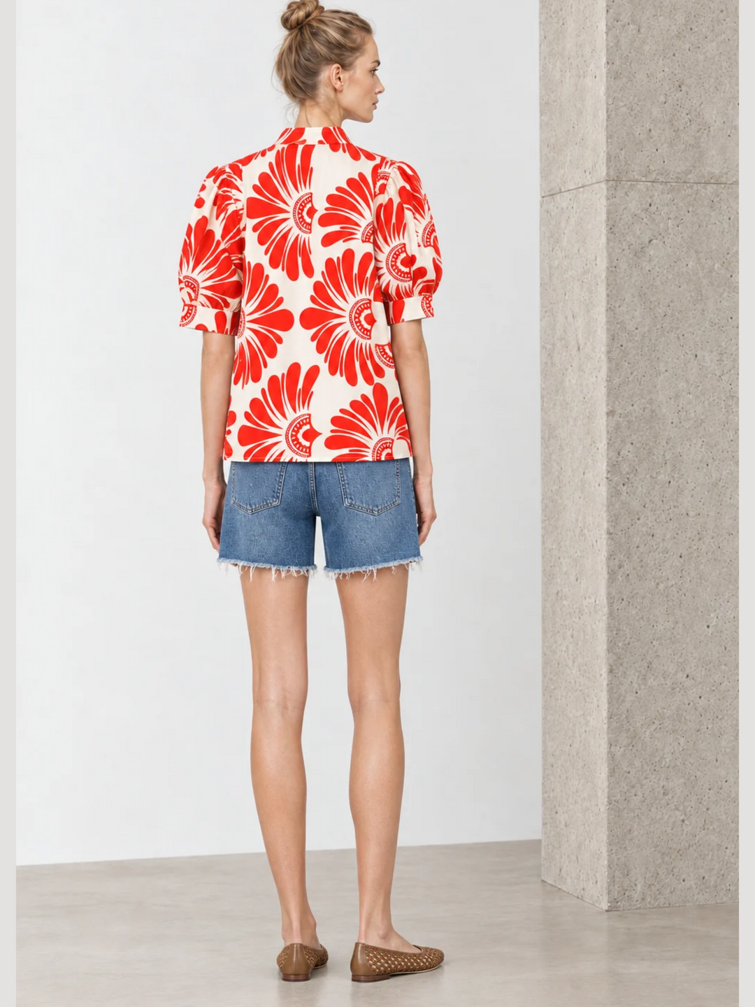 A woman stands with her back to the camera wearing a short-sleeve button-up blouse with a bold red and orange floral pattern on a cream background, paired with denim shorts and brown woven flats.