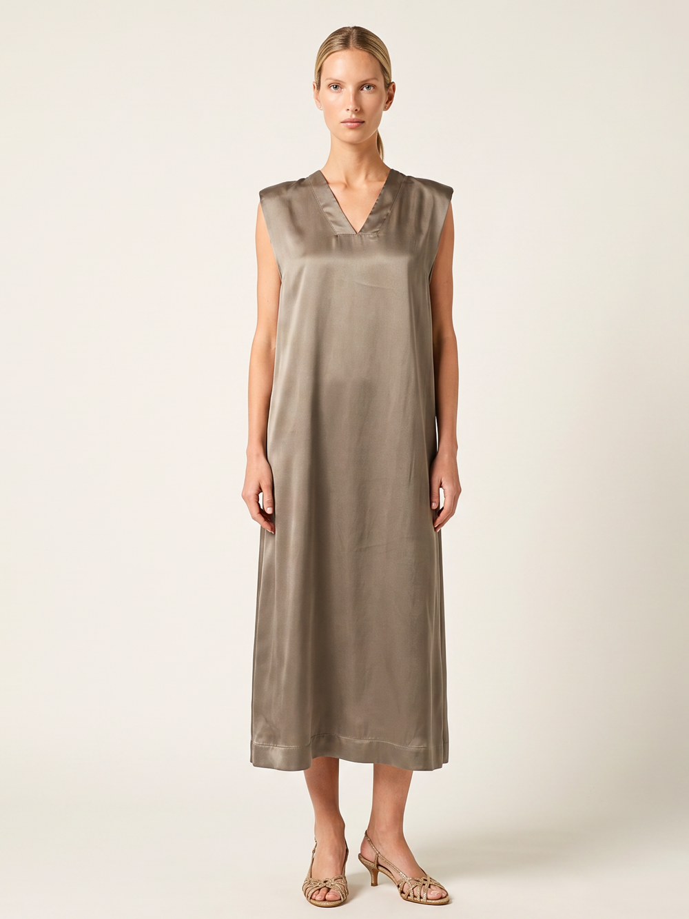 A woman modeling a sleeveless, V-neck midi dress in a shimmering taupe-brown satin fabric, standing against a plain light beige studio background.