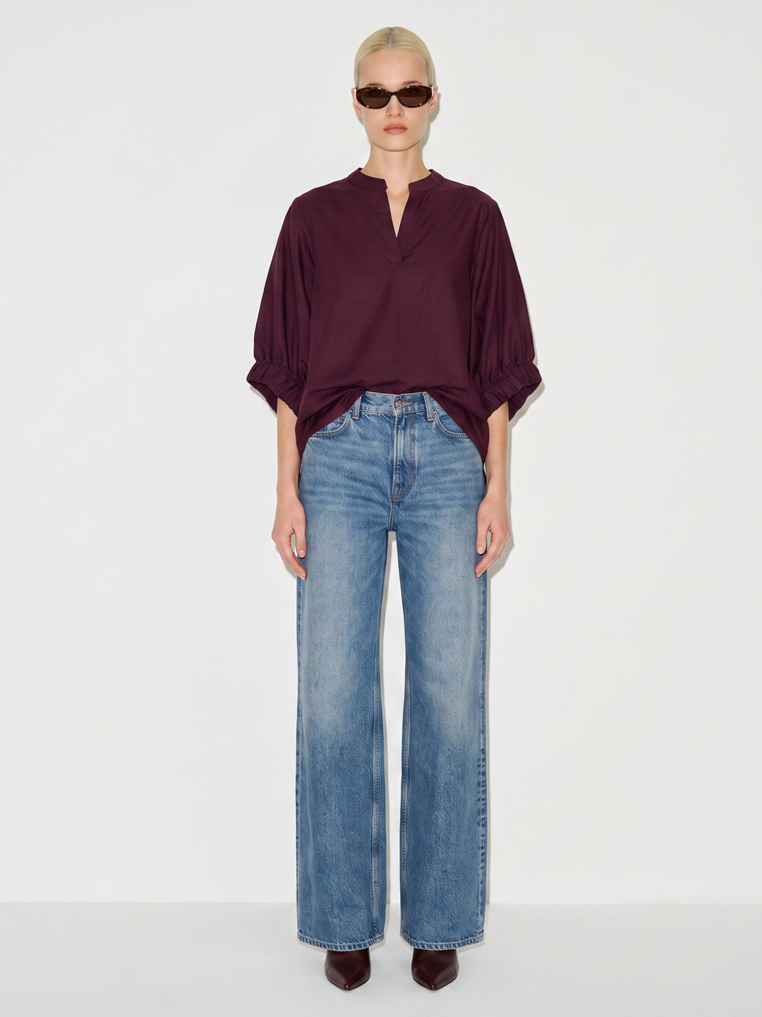 A model stands against a plain white studio background wearing a dark burgundy blouse with a split neckline and wide sleeves, paired with light-wash blue wide-leg jeans and dark brown pointed shoes.