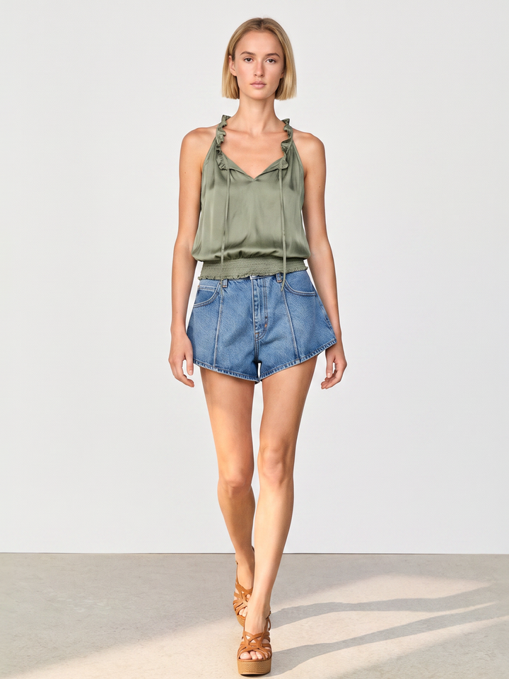 A model wearing an olive green satin halter top and blue denim shorts, walking against a light gray studio background.
